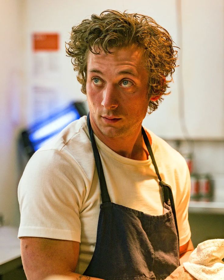 a man with curly hair wearing an apron and looking at the camera while holding food in his hands