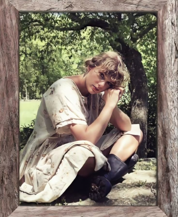 a woman sitting on the ground next to a tree and holding her hand near her face