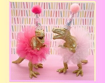 two small dinosaurs dressed in gold and pink tutues with pom poms