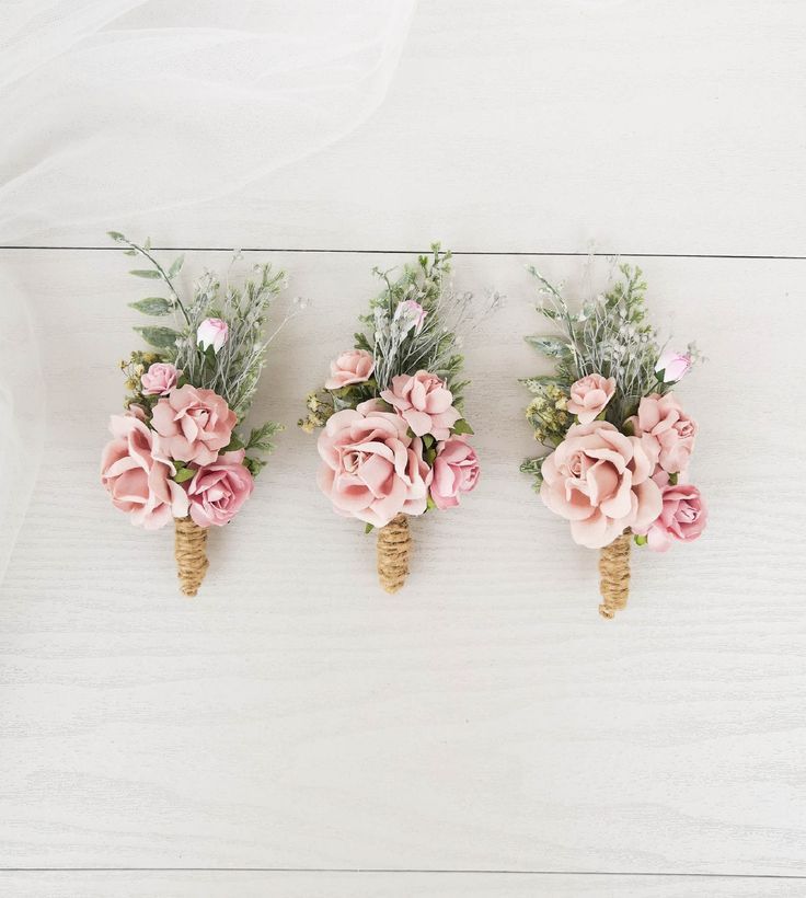 three boutonnieres with pink flowers and greenery in them on a white wooden surface