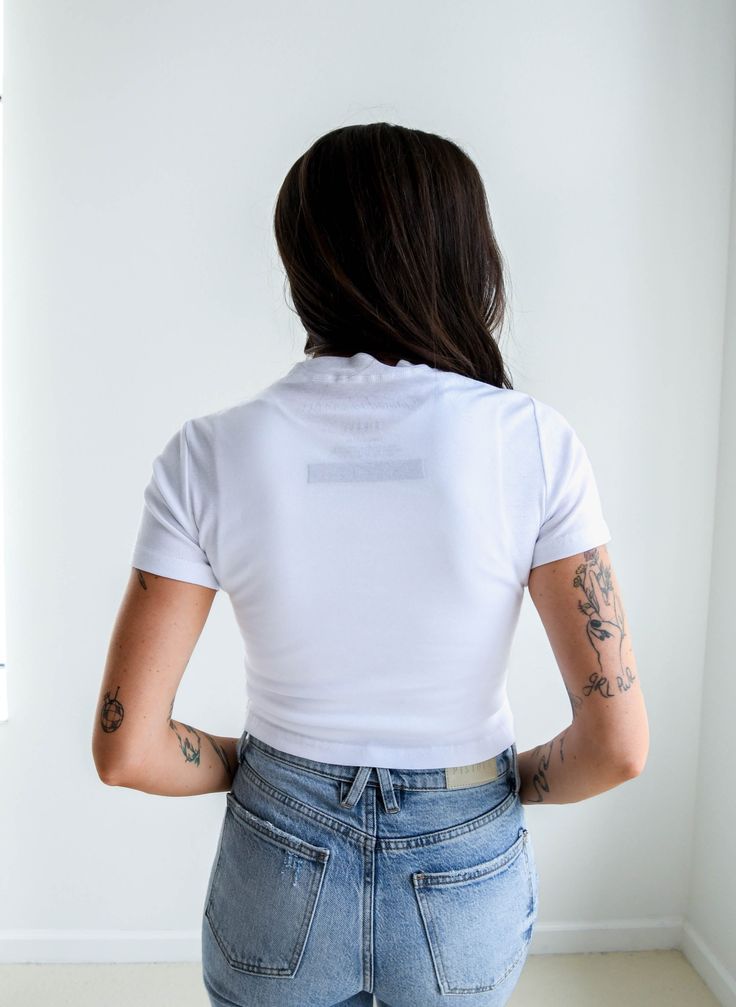 a woman with tattoos on her back standing in front of a window wearing jeans and a white t - shirt