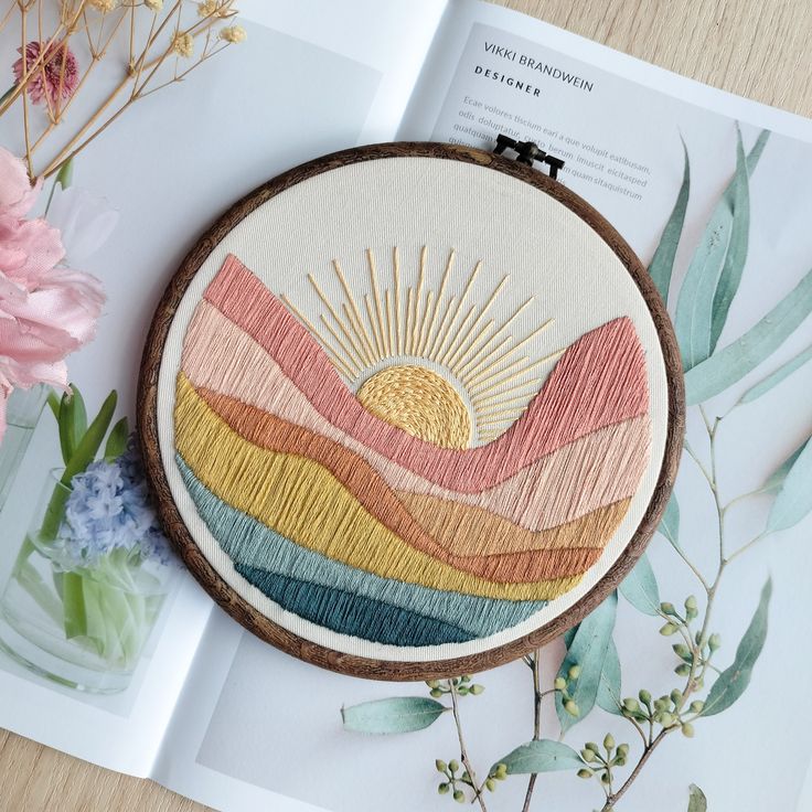 an embroidery project on a table next to flowers and a book with the sun in it