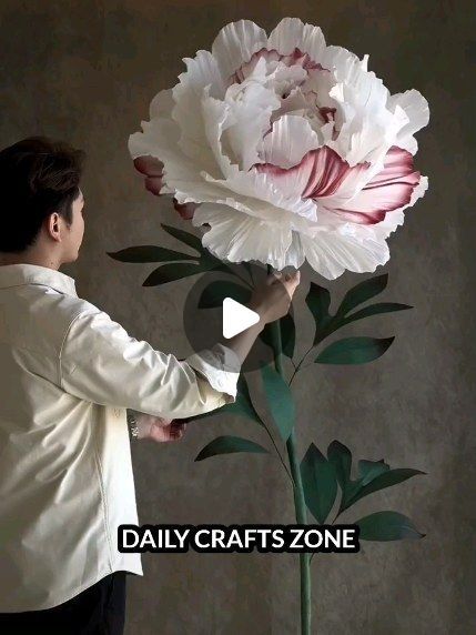 a man holding a large white flower in front of a gray wall with the words daily crafts zone written on it