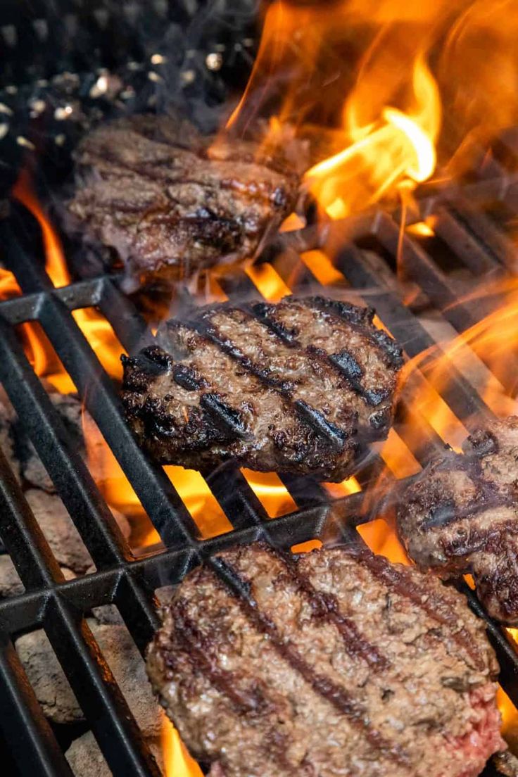 hamburgers and burger patties cooking on the grill with flames in the foreground