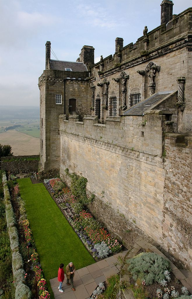 two people walking in front of an old castle like building with gardens and flowers on the lawn