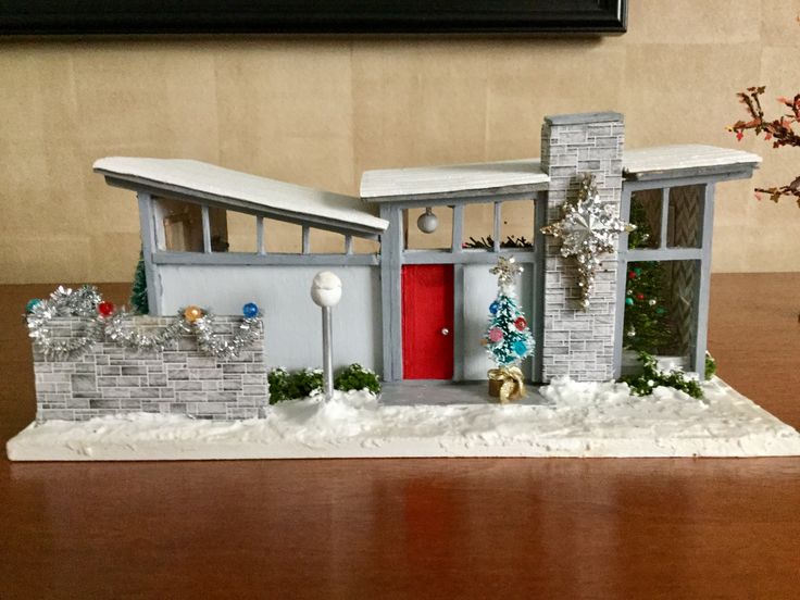 a model house with snow on the ground next to a christmas tree and potted plant