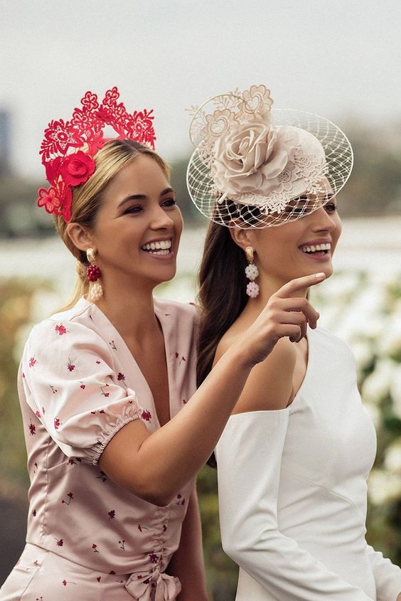 two beautiful women standing next to each other in front of flowers and trees, one wearing a hat with a flower on it