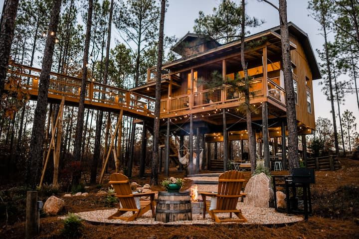 a house in the woods surrounded by trees with chairs around it and a fire pit