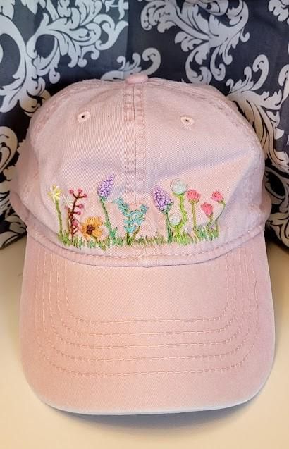 a pink baseball cap with embroidered flowers on the front and side, sitting against a patterned background