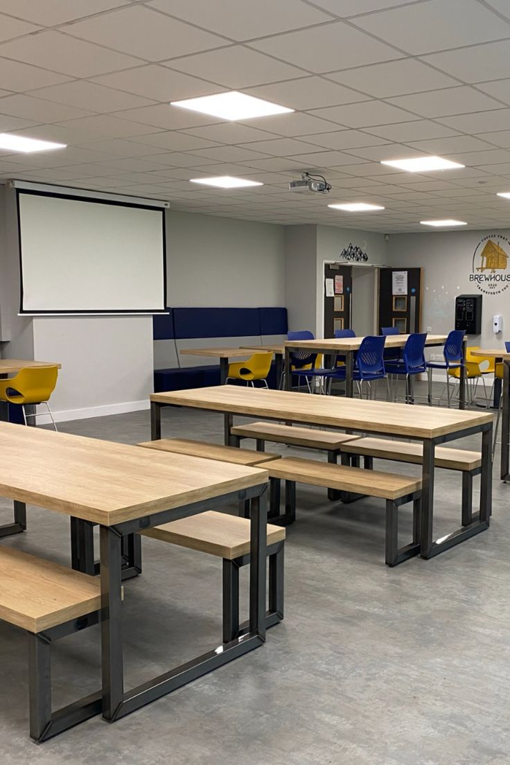 an empty classroom with tables and chairs in it's center area, while a projector screen is on the wall