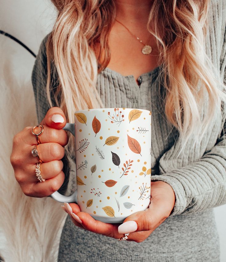 a woman holding a coffee mug with autumn leaves printed on the front and sides, in her hands