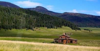 an old log cabin sits in the middle of a field with mountains in the background