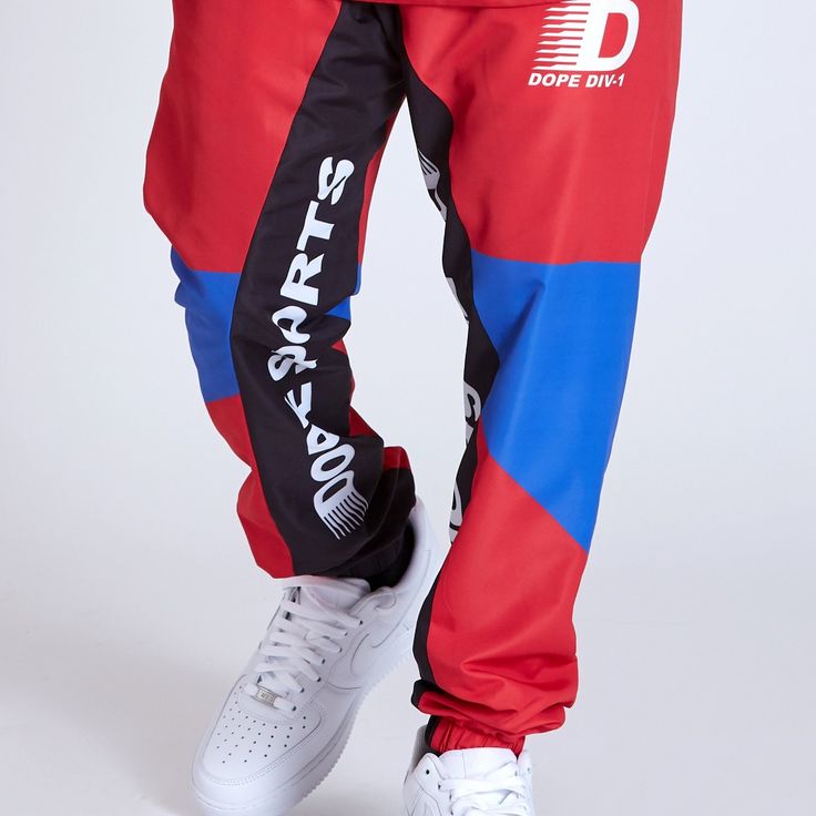 Division Track Pant By Dope. Polyester Windbreaker Jogger Pants With Paneled Construction. Self Elastic Drawstring Waist , Elastic Ankle Cuffs, Two Front Welt Hand Pockets And Single Welt Back Pocket With Woven Label Detail. Fine Mesh Lining. Style And Fit Classic Fit. Model Is 5'8" And Wears A Size M. Order Your Size For The Intended Fit. Sporty Multicolor Long Pants, Multicolor Straight Leg Bottoms For Streetwear, Multicolor Straight Leg Pants For Streetwear, Multicolor Wide Leg Streetwear Bottoms, Sporty Red Straight Leg Bottoms, Multicolor Wide Leg Bottoms For Streetwear, Multicolor Pants With Elastic Waistband For Streetwear, Straight Leg Bottoms With Elastic Side Panels For Streetwear, Blue Streetwear Bottoms With Elastic Side Panels