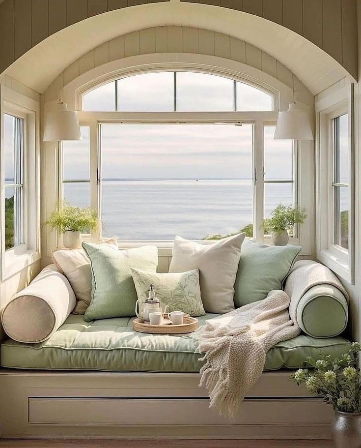 a window seat with pillows and blankets on it in front of an arched window overlooking the ocean