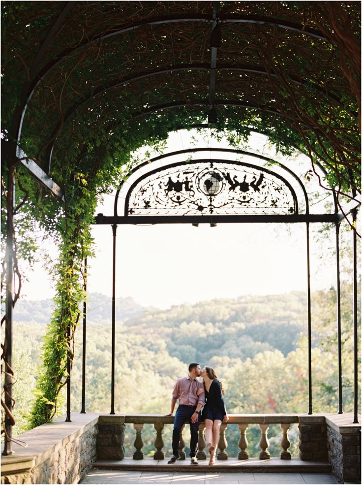 two people sitting on a bench under an arch with vines growing over it and mountains in the background