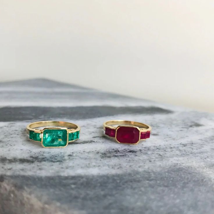 two gold rings with green and red stones on top of a stone slab in front of a white wall