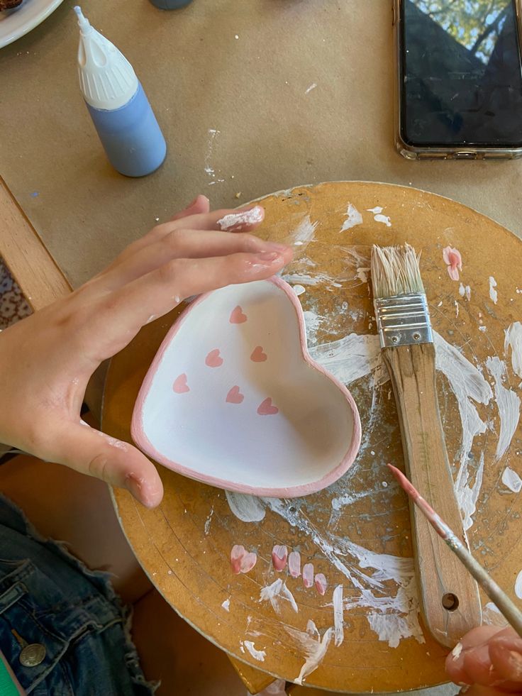 two hands holding a heart shaped bowl with paint on it and brushes in the other hand