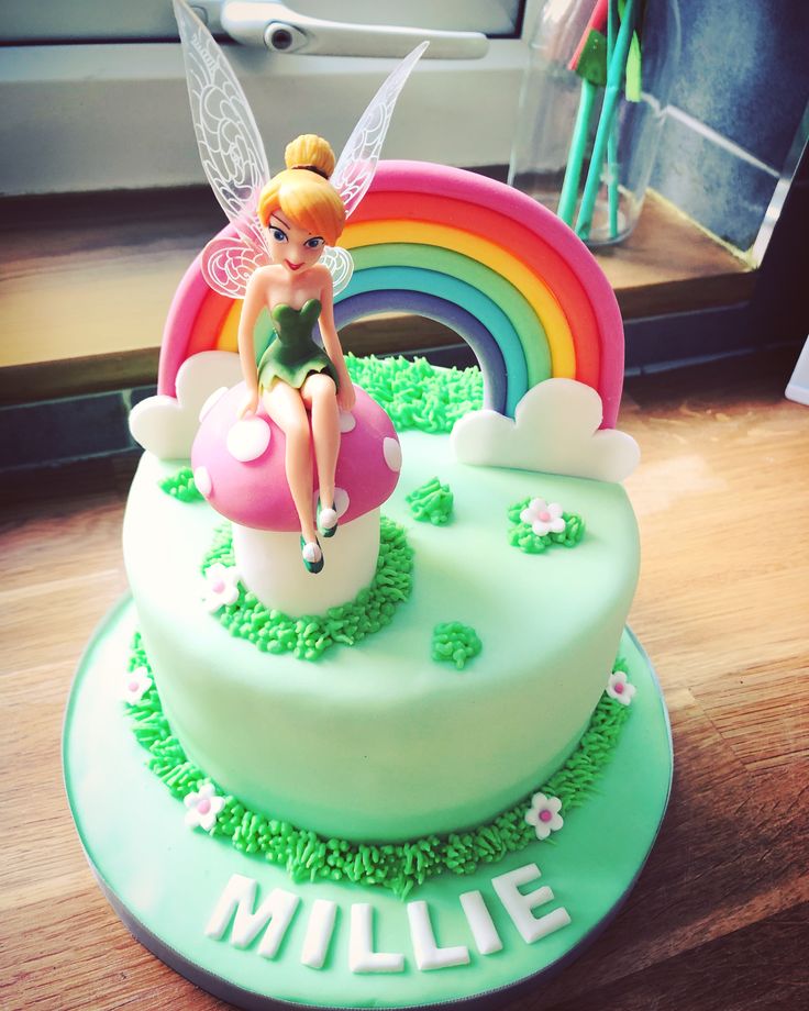 a green cake with a fairy sitting on top of it and rainbow in the background