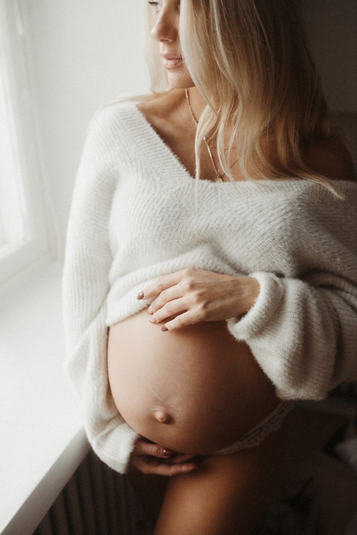 a pregnant woman wearing a white sweater standing next to a window with her hands on her belly