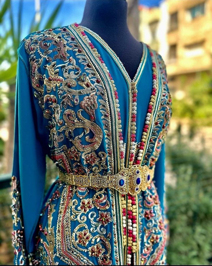 A Moroccan caftan in blue and gold with accents of red, meticulously crafted for performances and special events, is a stunning ensemble that seamlessly blends traditional elegance with contemporary flair. The deep blue fabric forms the foundation, creating a rich and regal backdrop for the intricate details. The golden embellishments, meticulously embroidered throughout the caftan, add a touch of opulence and sophistication. The design features elaborate patterns and motifs inspired by Moroccan Blue Festive Kaftan With Dabka, Blue Dabka Kaftan For Festive Occasions, Festive Blue Dabka Kaftan, Blue Bollywood Style Kaftan With Traditional Drape, Bollywood Style Gold Kaftan For Festive Occasions, Gold Bollywood Kaftan For Festive Occasions, Festive Blue Traditional Drape Kaftan, Blue Dabka Work Kaftan For Festivals, Gold Bollywood Kaftan