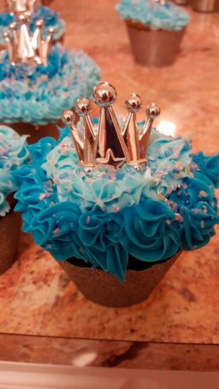 cupcakes with blue frosting and silver crowns on top are sitting on a table