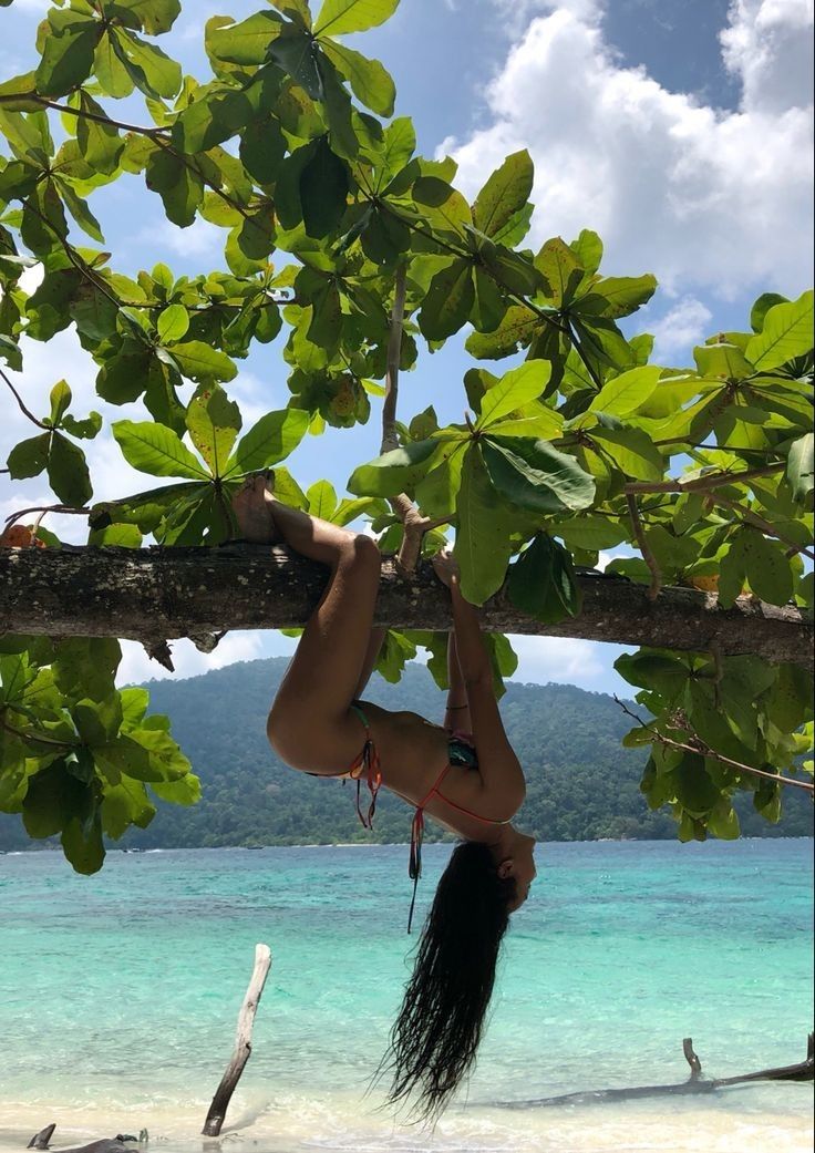 a woman hanging upside down from a tree branch on the beach with blue water in the background