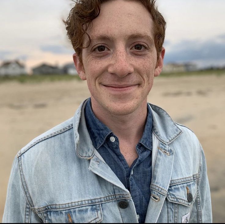 a young man with curly hair and blue shirt smiling at the camera while standing in front of an empty beach