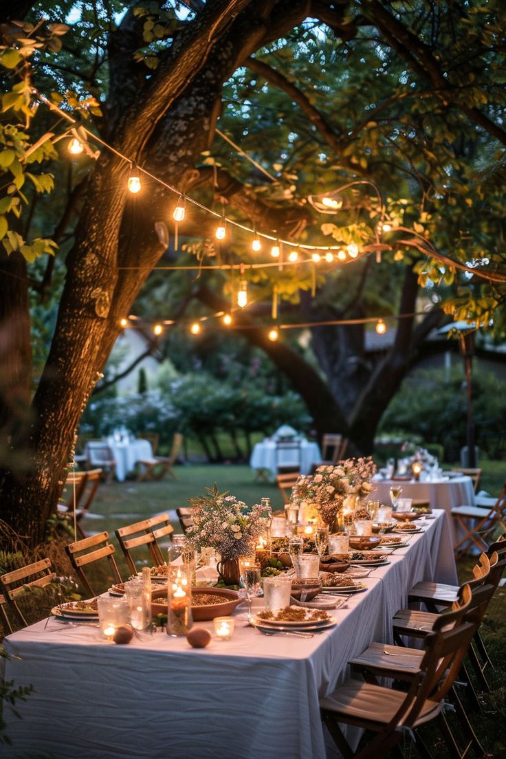 Outdoor table setup with white cloth, candles, and flower centerpieces under string lights hanging from tree branches in a garden during twilight. Garden Celebration Party Ideas, Barbecue Themed Party, Backyard Autumn Wedding, Backyard Garden Dinner Party, Outdoor Garden Party Ideas Decoration, Elegant Backyard Dinner Party, Summer Garden Dinner Party, Engagement Party Garden, Backyard 40th Birthday Party