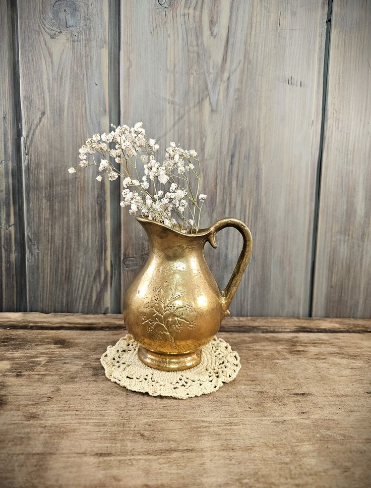 a gold pitcher with white flowers in it sitting on a doily next to a wooden wall