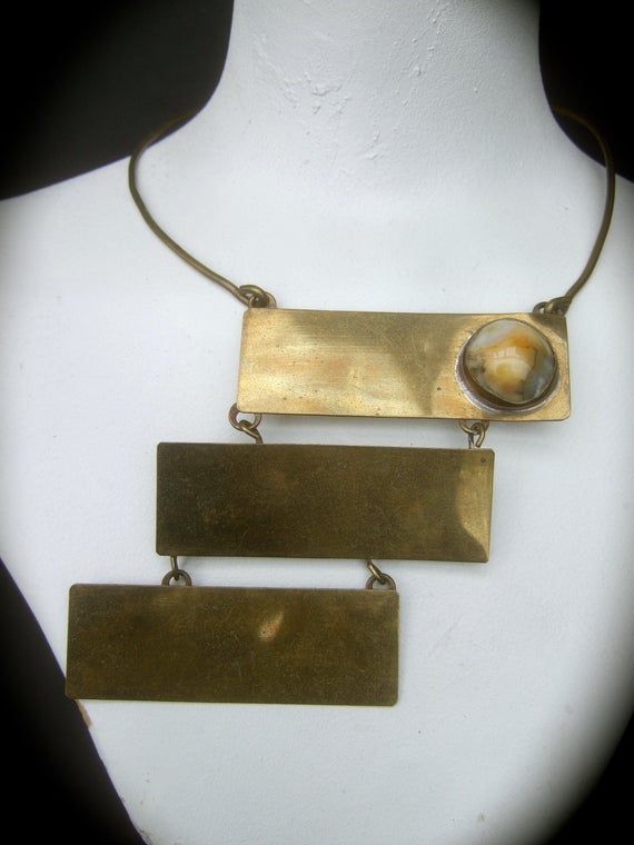 1970's Avant-garde brass metal articulated tile choker necklaceThe unique artisan choker necklace is designed with three large scale brass metal rectangular articulated tilesThe trio of brass metal tiles are suspended from a rigidwire band that wraps around the neck. One of the brassmetal rectangular tiles is adorned with a milky smooth glass cabochonThe edgy mod handmade artisan choker makesa very chic one of a kind accessory Measurements The rigid brass metal wire band interior circumferenceth Vintage Brass Rectangular Jewelry, Vintage Rectangular Brass Jewelry, Modernist Bronze Brass Jewelry, Antique Rectangular Brass Jewelry, Adjustable Rectangular Bronze Jewelry, Brutalist Brass Jewelry In Bronze, Rectangular Bronze Brass Jewelry, Brutalist Bronze Brass Jewelry, Vintage Square Metal Jewelry