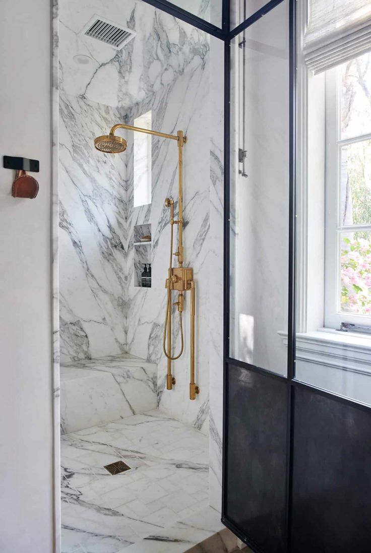 a bathroom with white marble walls and flooring, shower head, and window in the corner