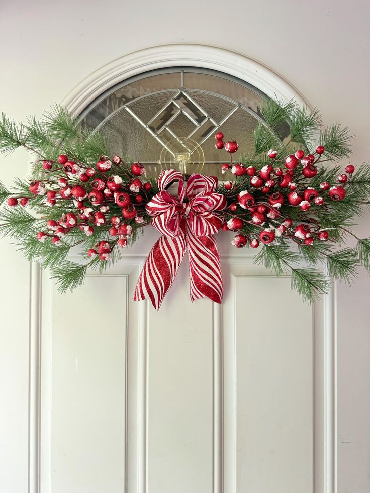 a christmas wreath with red and white ribbons hanging on the front door, decorated with greenery