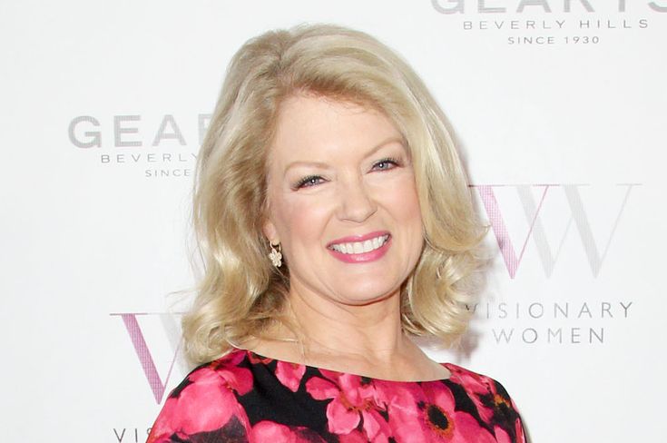 an older woman with blonde hair wearing a floral dress and smiling at the camera while standing in front of a white wall