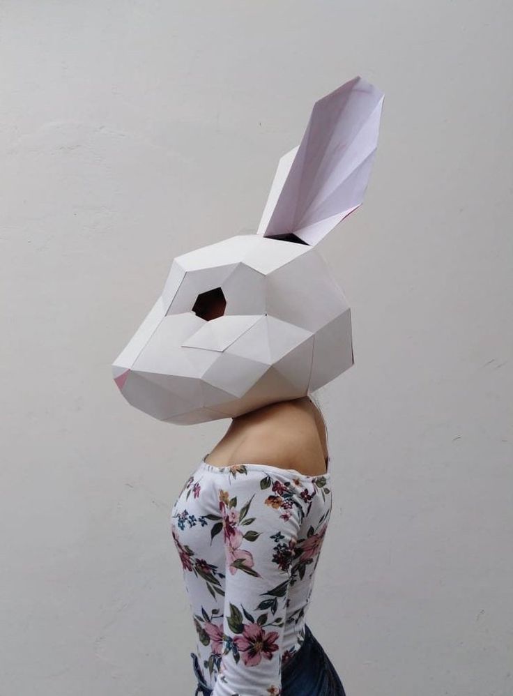 a woman wearing a paper bunny mask with flowers on it's head, standing in front of a white wall