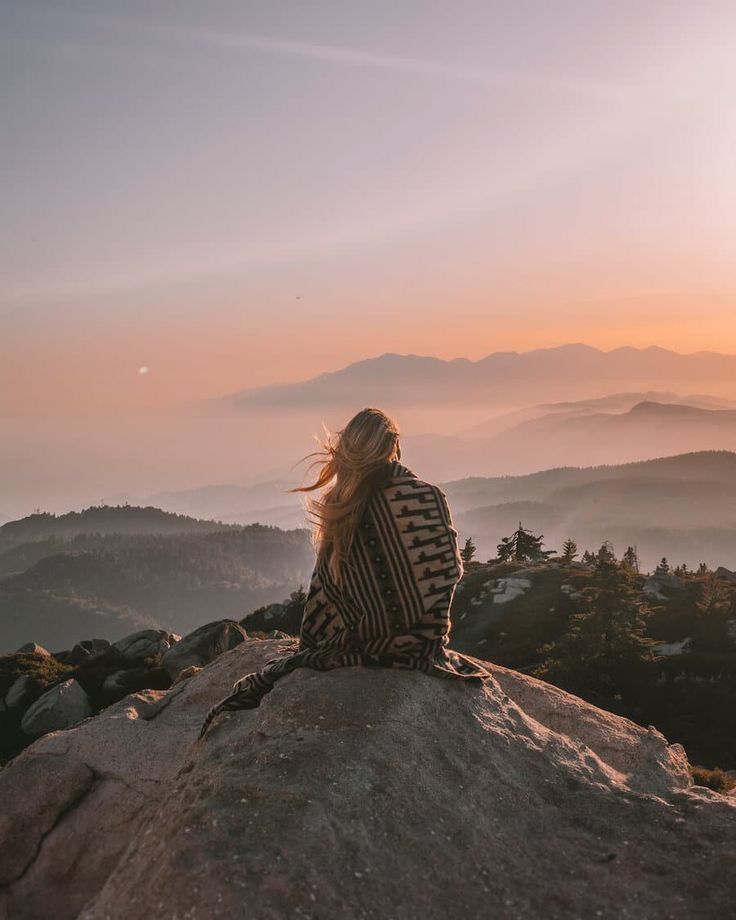 a woman sitting on top of a rock looking out at the mountains in the distance