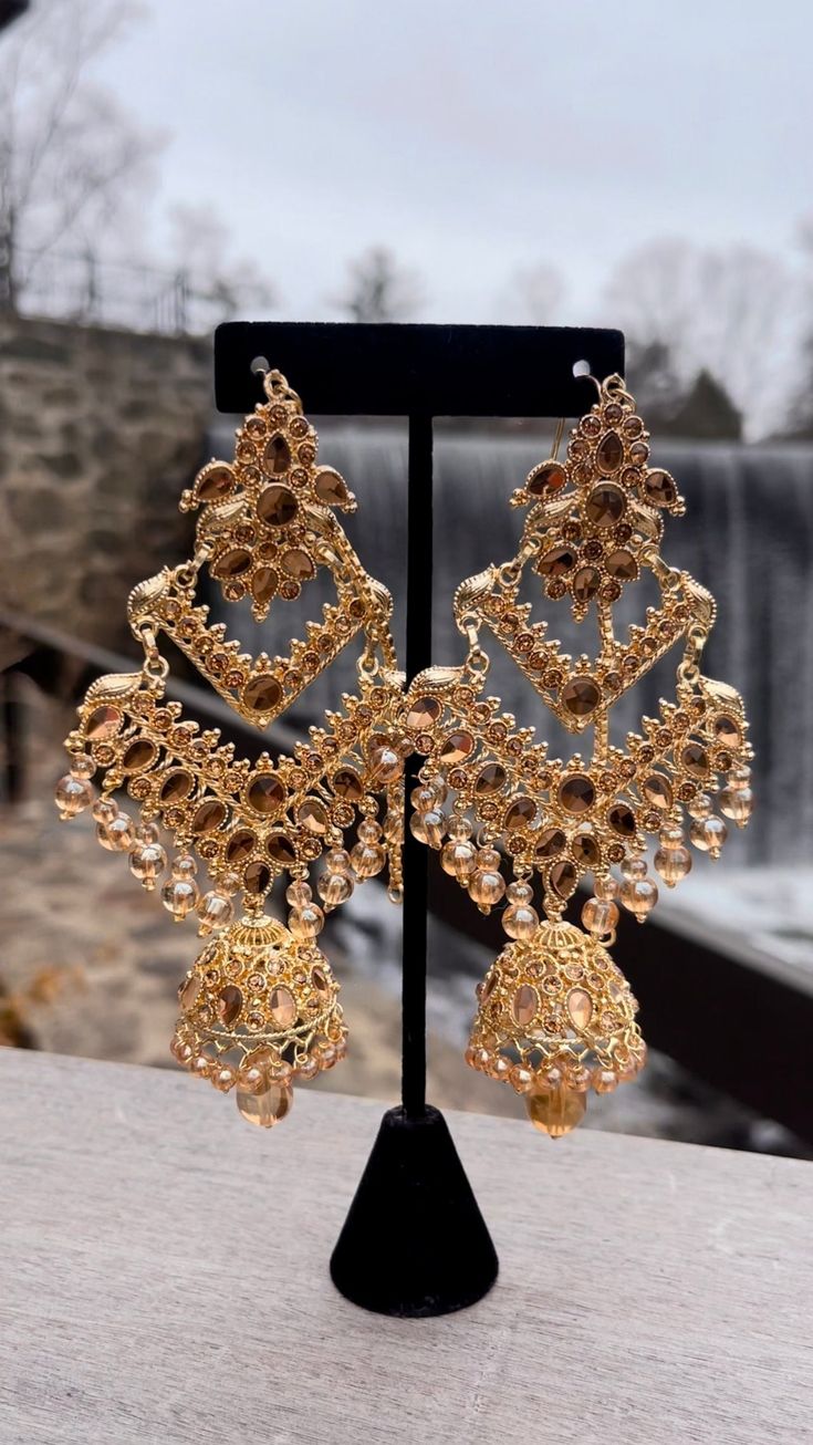 Below the top section, the earrings transition into a large, ornate diamond-shaped framework that is embellished with additional teardrop gemstones and intricate filigree work. The middle section is heavily decorated with small beads and dangling elements that add to the overall opulence. The earrings culminate in a grand jhumka, a traditional bell-shaped ornament encrusted with sparkling stones and detailed patterns. Dangling from the jhumka are multiple strands of beads, adding movement and a Kundan Chandbali, Small Beads, Diamond Shapes, Statement Earrings, Favorite Jewelry, Jewelry Earrings Dangle, Etsy Earrings, Dangle Drop Earrings, Dangle Earrings