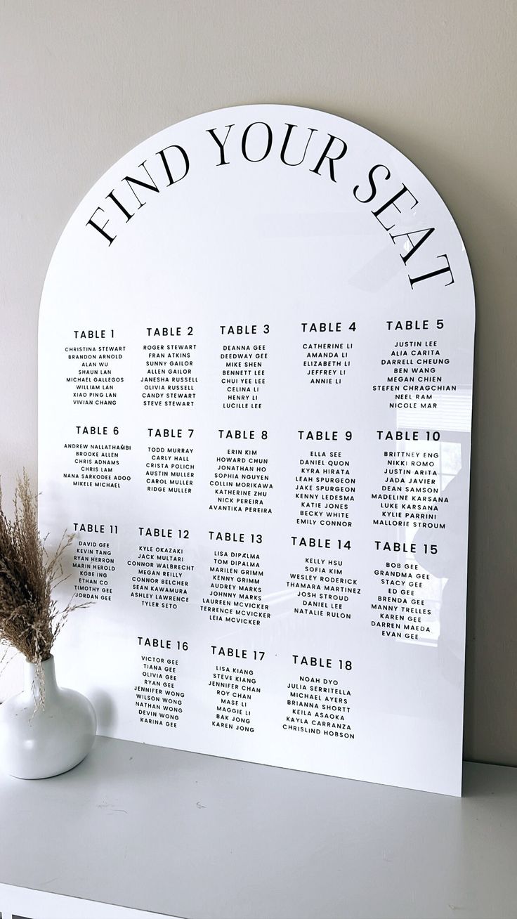 a table plan is displayed on a white shelf next to a vase with dried flowers