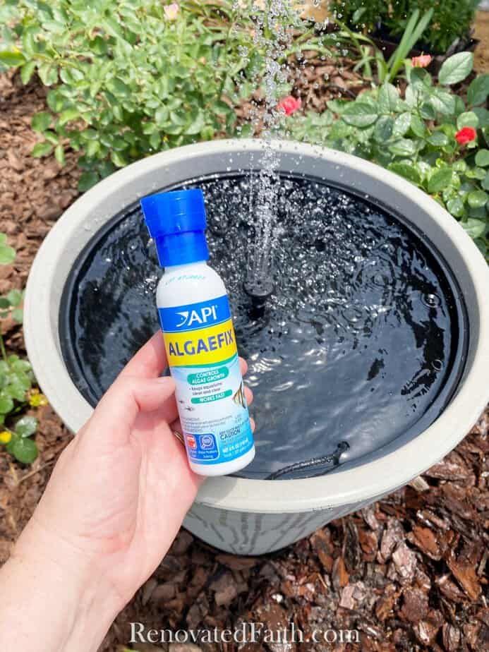 someone is holding a bottle of water in front of a fountain with plants and flowers around it