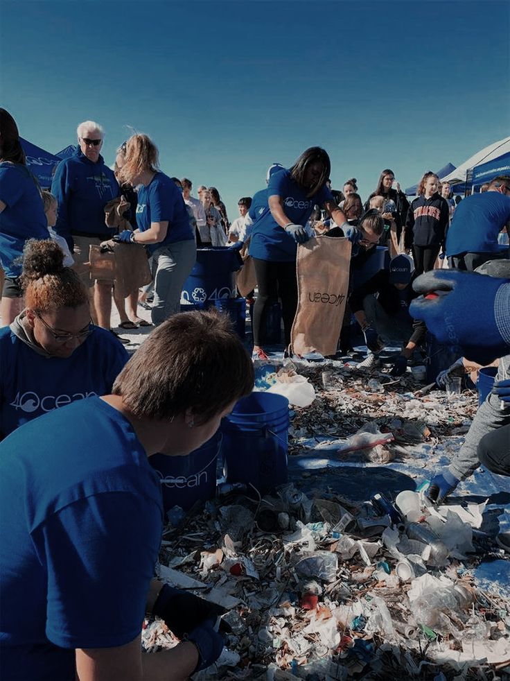 people in blue shirts are picking up trash at the beach while others stand around them