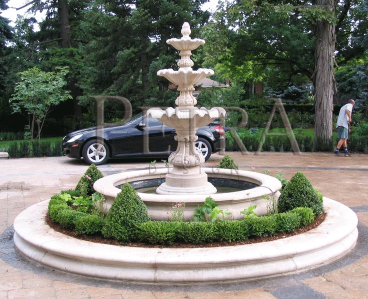 a car is parked next to a fountain in the middle of a park with trees and bushes