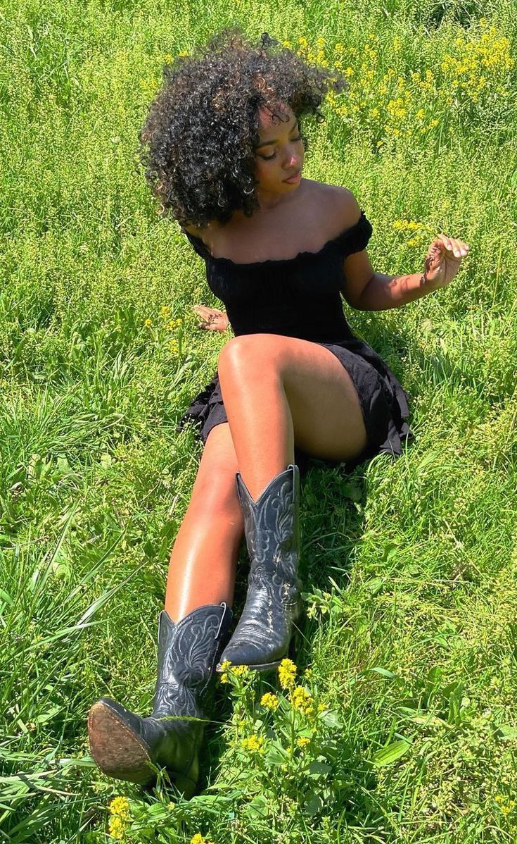 a woman sitting in the grass with her legs crossed and wearing cowboy boots, eating something