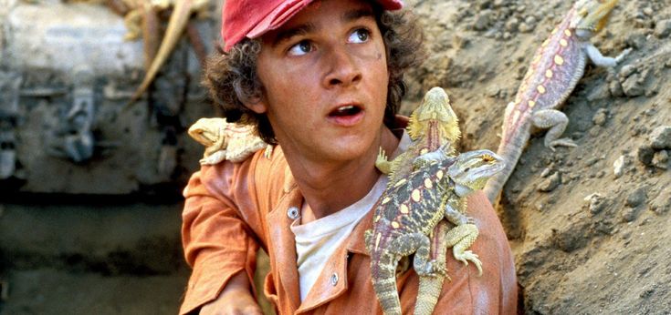 a young man in an orange jacket and red hat holding two lizards on his arm