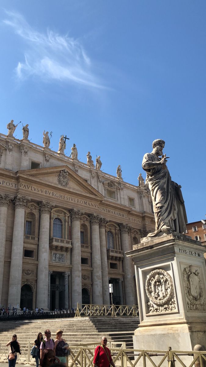 people are walking around in front of a large building with statues on the steps outside