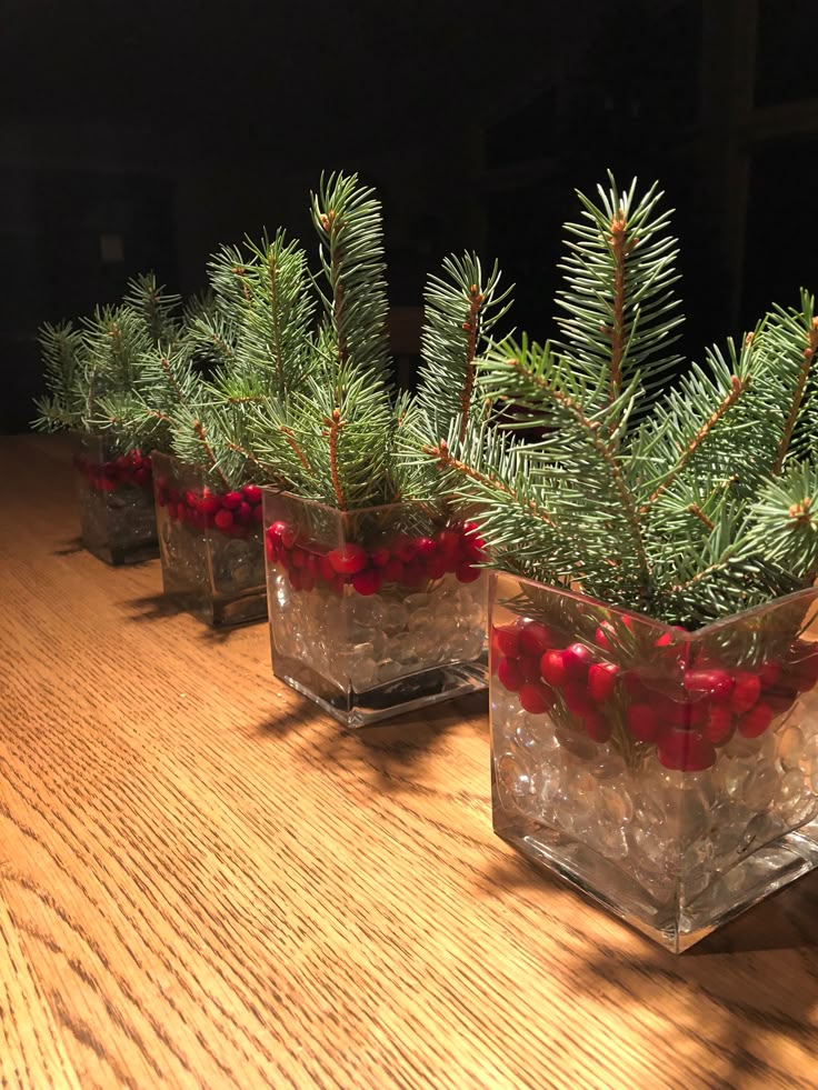 three clear glass vases filled with red berries and pine needles on a wooden table