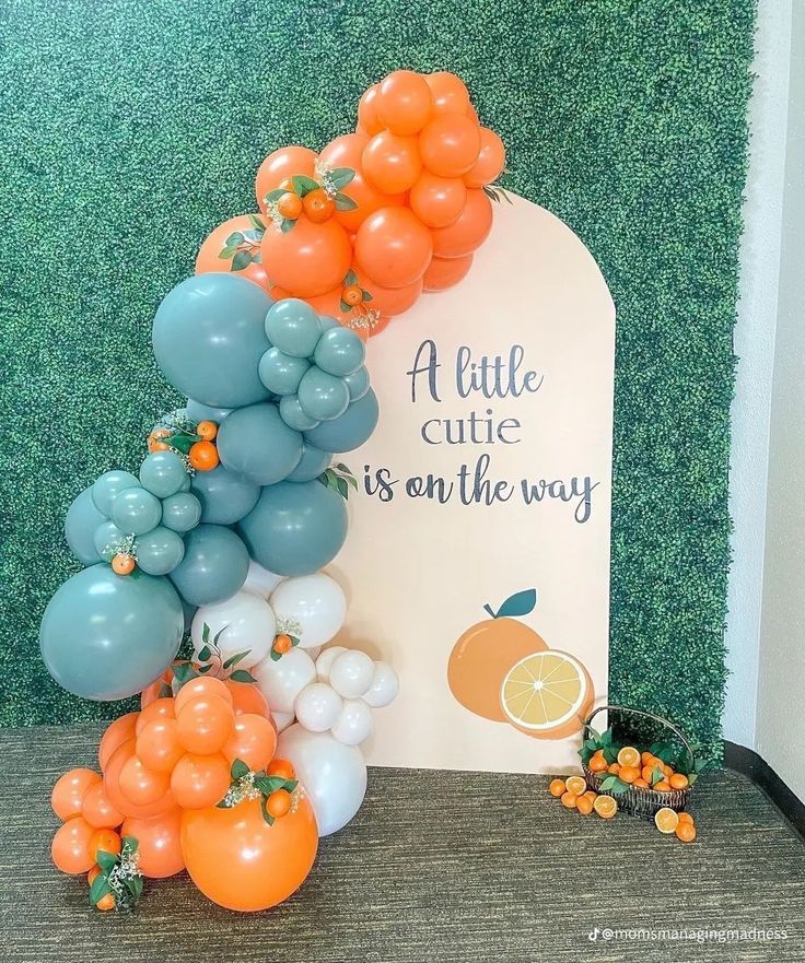 an arch made out of balloons and oranges on the grass with a sign that says a little cute is on the way