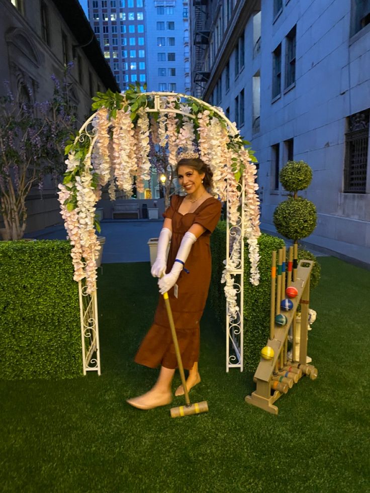 a woman in brown dress holding a broom and standing next to a white arch with flowers on it