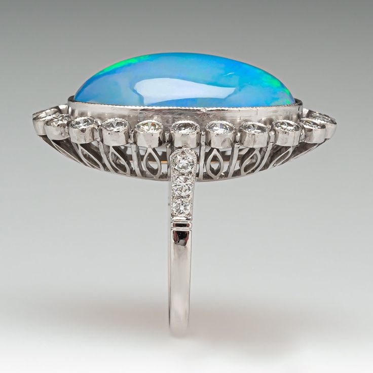 This gorgeous ring is centered with one (1) oval cabochon cut natural crystal opal and set into a bezel setting. The opal is bordered with twenty-two (22), bezel set, round brilliant cut diamonds, The top face of the shank is accented with three (3), bead set, round brilliant cut diamonds on each side. The gemstones are bordered with milgrain edging. The ring measures 29.1mm at the top, rises 10.6mm above the finger, tapering to 1.8mm wide and 1.1mm thick at the base of the shank. This ring is currently a size 6. Round Cabochon Opal Anniversary Ring, Oval Opal Ring With Bezel Setting For Formal Occasions, Cabochon Opal Ring For Anniversary, Round Cabochon Opal Ring For Anniversary, Formal Oval Cabochon Opal Ring, Oval Opal Ring With Polished Finish In Fine Jewelry, Heirloom Opal Ring With Bezel Setting For Formal Events, Heirloom Bezel Set Opal Ring For Formal Occasions, Heirloom Style Bezel Set Opal Ring For Formal Occasions