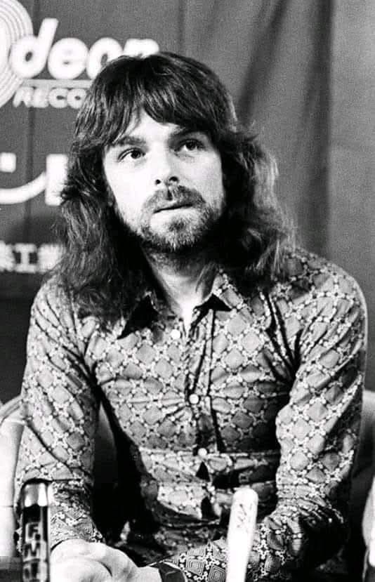 a man with long hair sitting at a table in front of a microphone and looking off to the side