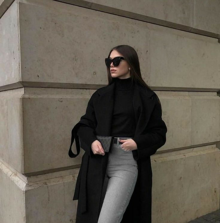 a woman standing in front of a building wearing sunglasses and a black coat with her hands on her hips