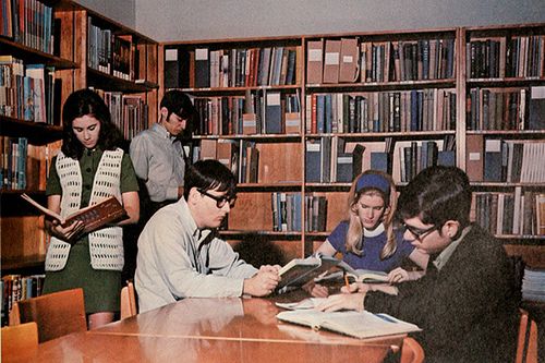 some people are sitting at a table in front of bookshelves and one person is holding an open book